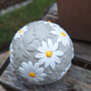 Könnte beinhalten: Eine kugelförmige Gartenkugel aus Beton mit einem erhabenen, geprägten Gänseblümchenmuster. Die Blumen sind mit weißen Blütenblättern und leuchtend gelben Mitten bemalt und heben sich von einer glatten, hellgrauen Betonoberfläche mit weiteren unbemalten floralen Texturen ab.