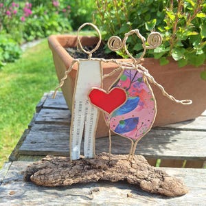 Könnte beinhalten: Eine handgefertigte Drahtskulptur von zwei Figuren, die ein rotes Herz halten. Eine Figur hat einen Papierkörper mit Text, die andere ein rosa Blumenkleid. Die Skulptur ist auf einem Stück Treibholz montiert, mit verschwommenem grünem Hintergrund.