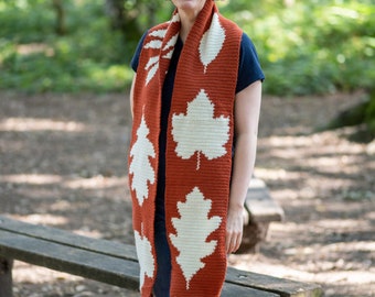 May include: A rust-colored crochet scarf with white leaf designs. The scarf has a fringed edge with white and rust-colored tassels. The scarf is worn by a person in a forest setting.