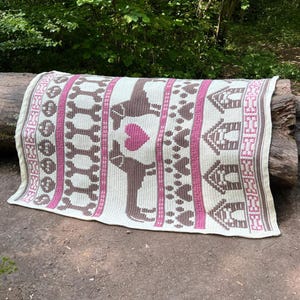 Puede incluir: Una manta de punto color crema con un patrón repetido de perros salchicha marrones, corazones rosas y casetas para perros. La manta está colocada sobre un tronco en un entorno natural al aire libre. El diseño incluye huesos de perro y huellas de patas.