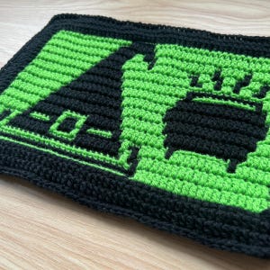 May include: A rectangular crochet piece featuring a black border and a vibrant green background. The design displays a black silhouette of a pointed witch hat on the left and a black cauldron with rising steam on the right, all crafted in yarn.