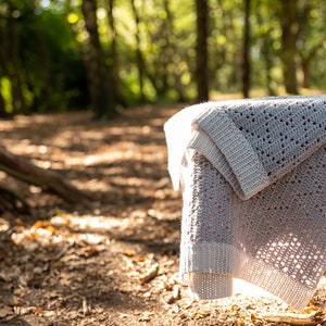 May include: A light grey crocheted blanket with a delicate open stitch pattern, draped over a wooden bench in a woodland setting.