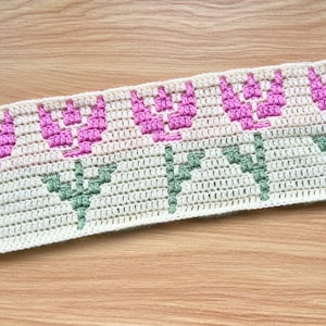 May include: Crocheted fabric strip featuring a repeating floral pattern. The design includes pink tulip-like flowers with green stems and leaves, set against a cream-colored background. The craftwork is displayed on a wooden surface.
