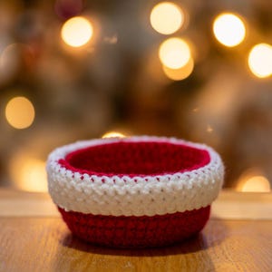 May include: A small, red and white crocheted bowl. The bowl is empty and sitting on a wooden surface.