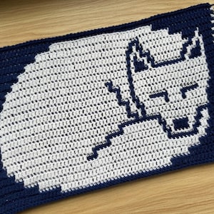 Puede incluir: Una manta de ganchillo hecha a mano con un diseño de fantasma pixelado en blanco sobre un fondo azul marino. La manta es rectangular con un borde azul marino. El diseño del fantasma es una representación simple y en bloques.