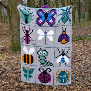 May include: A gray, handmade crochet blanket featuring a grid of colorful insect designs. The blanket showcases a variety of bugs, including a butterfly, bee, and ladybug, each in a pixelated style. The blanket is held up in a natural outdoor setting.