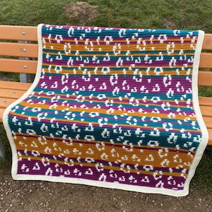 May include: A vibrant, hand-crocheted blanket with a repeating pattern. The blanket showcases stripes of teal, mustard, and purple, with white leopard-style spots. The edges are finished with a cream border. This cosy blanket is ideal for adding a touch of handmade charm to any room.