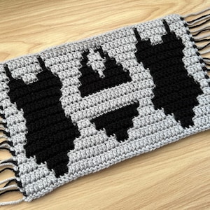 May include: Handmade crochet item featuring a geometric design in black on a gray background. The piece has a rectangular shape with fringe on the ends. The design appears to be a stylized cityscape or architectural pattern.