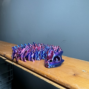 May include: A 3D printed, iridescent, purple and pink dragon figurine. The dragon is curled up in a sleeping position with its head resting on the wooden surface.