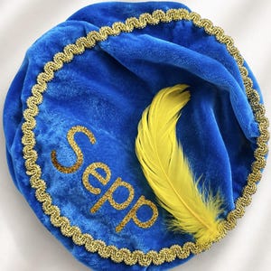 Op de afbeelding: Een blauwe fluwelen hoed met een gouden gevlochten rand langs de cirkelvormige omtrek. De naam Sepp is in gouden glitterletters op de voorkant geborduurd, vergezeld van een enkele felgele veer bevestigd aan de zijkant van de zachte stoffen kroon.