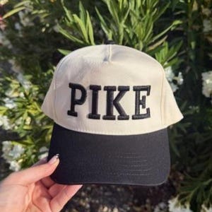 May include: A cream-colored baseball cap with a black brim. The word "PIKE" is embroidered in bold, black letters across the front of the cap. The cap is set against a backdrop of green foliage and white flowers.