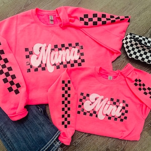 May include: Two pink sweatshirts with black and white checkered sleeves. The larger sweatshirt has the word "Mama" in white on a black and white checkered background. The smaller sweatshirt has the word "Mini" in white on a black and white checkered background.