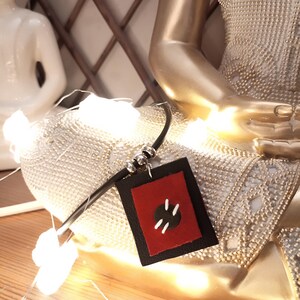 May include: A black leather necklace with a red and black square pendant. The pendant has a black circle with white stitching.