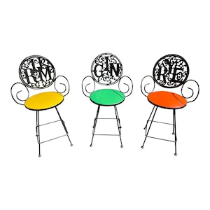May include: Three black metal bar stools with ornate scrollwork and a circular back. Each stool has a different colored seat cushion: yellow, green, and orange. The back of each stool features a different word: "I'm", "Gin", and "Rye".