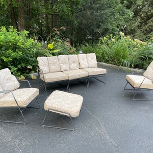May include: A mid-century modern patio furniture set with a sofa, two chairs, and an ottoman. The furniture is made of black metal frames with beige cushions. The cushions have a tufted design.