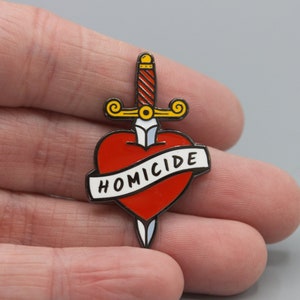 Może przedstawiać: Czerwone serce przebite srebrnym sztyletem i białym banerem z napisem "HOMICIDE" czarnymi literami. Sztylet ma żółtą rękojeść i srebrne ostrze.