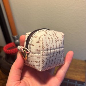 May include: A small, white fabric pouch with a zipper closure and a gold ring. The pouch is covered in a brown script pattern.