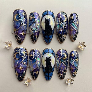 Peut inclure: Un ensemble de dix faux ongles en forme d'amande arborant un thème céleste avec des dégradés de bleu et de violet, des étoiles en feuille d'or, des croissants de lune et des silhouettes de chats noirs sur fond de pleine lune, le tout souligné de lignes dorées.