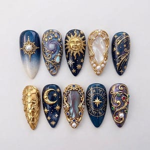 Op de afbeelding: Een set van tien amandelvormige press-on nagels met hemelse thema's. Ontwerpen bevatten gouden zonnen, manen, sterren, dierenriemwielen en sterrenstelsels op donkerblauwe en paarse achtergronden, geaccentueerd met parels, strass en goudfiligraan.