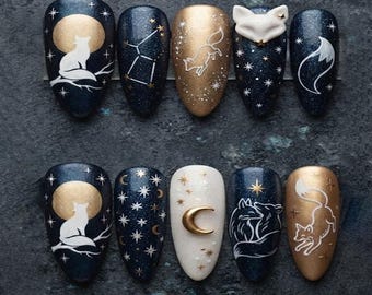 Puede incluir: Un juego de diez uñas postizas con diseños celestiales y de zorros. Las uñas muestran colores azul oscuro, dorado y blanco con estrellas, lunas, constelaciones y siluetas de zorros pintadas, incluyendo una uña con una cabeza de zorro en 3D.