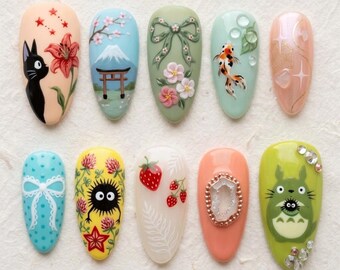 Op de afbeelding: Een set van tien amandelvormige kunstnagels met diverse handgeschilderde ontwerpen, waaronder een zwarte kat, de berg Fuji, een groene strik, een goudvis, sterpatronen, een blauwe strik, een roetwezentje, aardbeien, een kristal en een bosgeest.