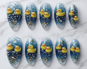 Op de afbeelding: Een set van tien kunstnagels met een blauwe gradiëntachtergrond, voorzien van verhoogde gele badeendjes, transparante bubbelaccenten en gouden glitterspikkels. Elke nagel heeft een puntige amandelvorm en een glanzende afwerking.