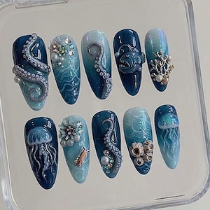 May include: A set of ten press-on nails featuring an ocean theme with deep blue and teal gradients. Designs include 3D octopus tentacles, a glowing anglerfish, jellyfish, coral, barnacles, and small pearls set against swirling water patterns.