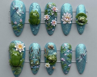 Puede incluir: Un juego de diez uñas postizas en forma de almendra con una base de purpurina azul claro. Los diseños incluyen ranas verdes esculpidas en 3D, nenúfares blancos y rosas, hojas de loto, libélulas y un pequeño caracol con temática de estanque.