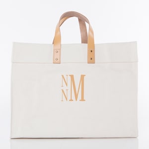 May include: Cream-colored tote bag with tan leather handles and accents. The bag features the monogram "NM" in a matching tan color. The handles are attached with rivets. The bag is rectangular and appears to be made of canvas.