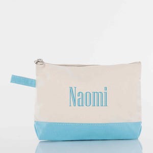 May include: A canvas cosmetic bag with a light blue base and handle. The upper portion is off-white, with the name "Naomi" printed in light blue. The bag has a silver zipper closure.