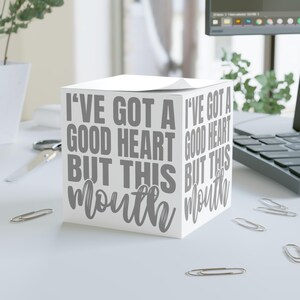 May include: A white cube-shaped notepad with the text "I'VE GOT A GOOD HEART BUT THIS MOUTH" in gray lettering. The notepad is on a white desk with scattered paper clips.
