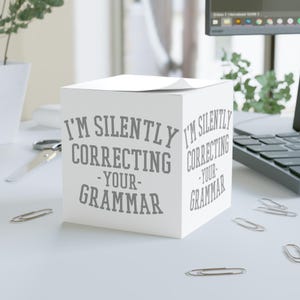 May include: A white cube-shaped notepad with the text "I'M SILENTLY CORRECTING -YOUR- GRAMMAR" printed in gray. The notepad is on a white desk with paper clips and a laptop in the background.