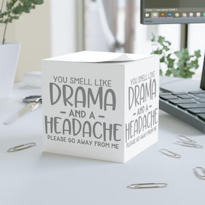 May include: A white cube-shaped notepad with the text "YOU SMELL LIKE DRAMA - AND A - HEADACHE PLEASE GO AWAY FROM ME" printed in gray. The notepad is on a white desk with paper clips and a laptop in the background.