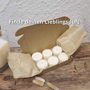 Könnte beinhalten: Eine offene Pappschachtel mit sechs weißen Teelichtkerzen mit silbernen Rändern. Die Kerzen sind in der Schachtel auf einer Holzoberfläche angeordnet. Der Text "Finde deinen Lieblingsduft" ist oben im Bild zu sehen.