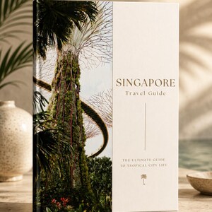 Könnte beinhalten: Ein Reiseführer mit dem Titel "SINGAPORE Travel Guide" mit einem Cover, das eine grüne Landschaft mit hohen Bäumen und einem geschwungenen Steg zeigt. Das Buch liegt auf einer hellen Oberfläche.