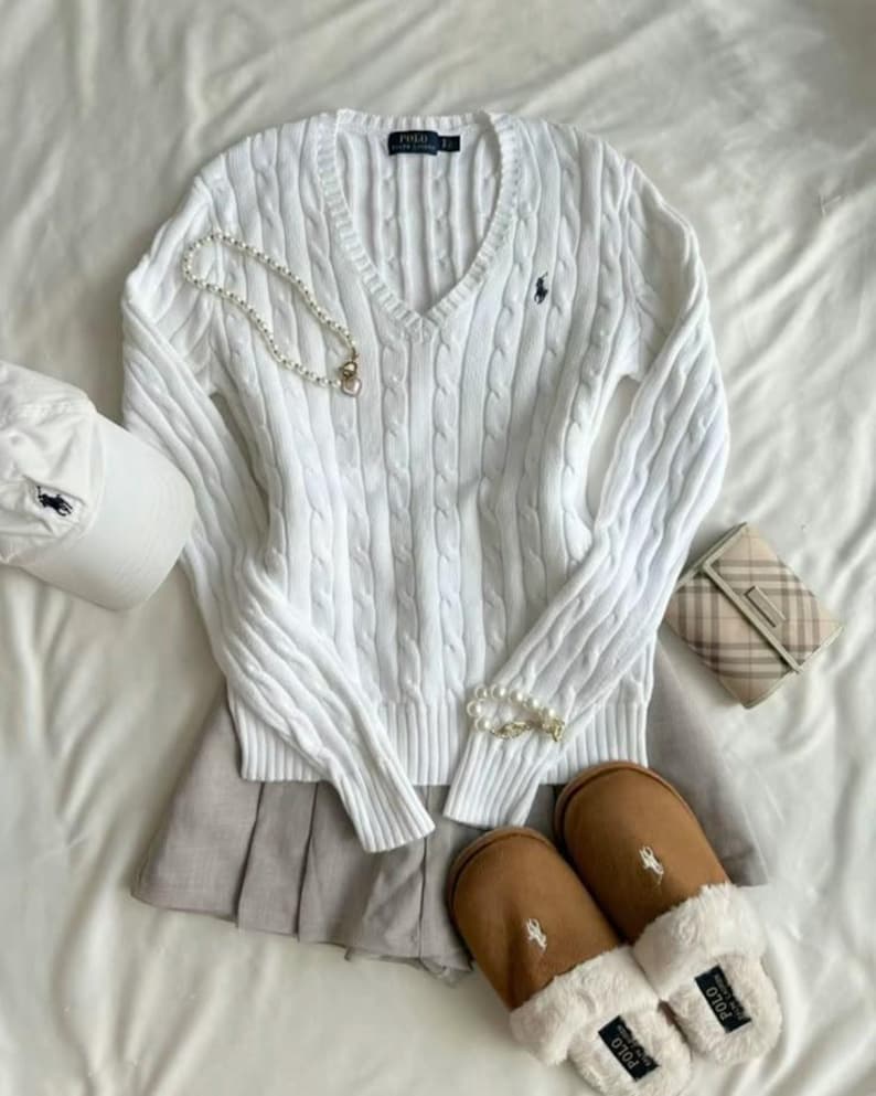 Pu&ograve; includere: Un maglione bianco a trecce con scollo a V, un berretto da baseball bianco e pantofole marroni con finiture in ecopelliccia bianca sono disposti su una superficie bianca. Sono presenti anche una collana di perle e una piccola borsa.