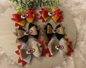 May include: Six glittery hair bows in red, gold, black, and silver. Each bow features a team logo in the center. The bows are arranged on a light wooden surface, with greenery in the background. These bows are perfect for game day.