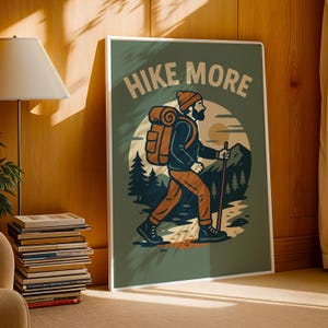 May include: A framed art print with the words "HIKE MORE" at the top. The illustration depicts a hiker with a backpack and walking stick, traversing a mountain landscape. The color palette includes shades of green, brown, and orange.