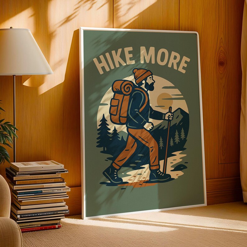 May include: A framed art print with the words "HIKE MORE" at the top. The illustration depicts a hiker with a backpack and walking stick, traversing a mountain landscape. The color palette includes shades of green, brown, and orange.