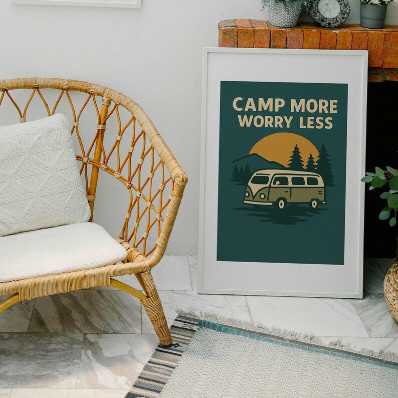May include: A framed art print with the text "CAMP MORE WORRY LESS" above a graphic of a vintage camper van, trees, and a setting sun. The print is in a white frame and is displayed next to a wicker chair and a patterned rug.