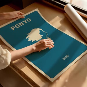 Puede incluir: Un p&oacute;ster turquesa con la palabra "PONYO" en blanco, una silueta blanca de un personaje y el a&ntilde;o "2008". Una persona sostiene el p&oacute;ster. El p&oacute;ster est&aacute; sobre una superficie de madera.