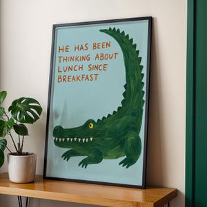 Könnte beinhalten: Ein gerahmter Kunstdruck mit einer Illustration eines grünen Alligators auf hellblauem Hintergrund. Der orangefarbene Text lautet: "HE HAS BEEN THINKING ABOUT LUNCH SINCE BREAKFAST." Der Alligator hat ein gelbes Auge und weiße Zähne.