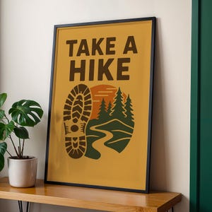 May include: Framed poster with the text "TAKE A HIKE" in brown letters. The poster features a brown boot print, a stylized mountain scene with green trees, a winding path, and a setting sun, all on a mustard yellow background.