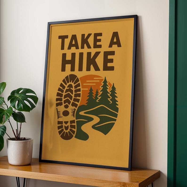 May include: Framed poster with the text "TAKE A HIKE" in brown letters. The poster features a brown boot print, a stylized mountain scene with green trees, a winding path, and a setting sun, all on a mustard yellow background.