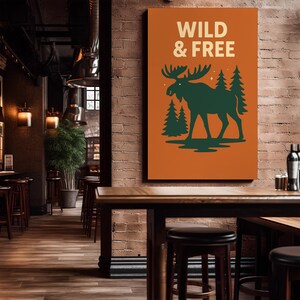 May include: An orange rectangular wall art piece with the words "WILD & FREE" in cream-coloured text. A dark green moose silhouette stands in front of two green pine trees. The art is displayed in a rustic bar setting.