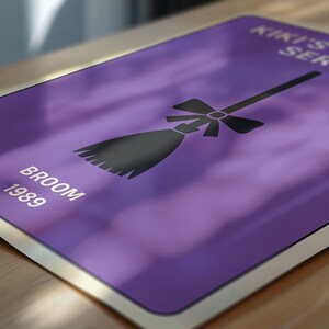 Puede incluir: Un p&oacute;ster morado con un gr&aacute;fico de escoba negra y el texto "Kiki's Service" y "Broom 1989". La escoba est&aacute; representada con un lazo. El p&oacute;ster tiene un borde blanco y est&aacute; sobre una superficie de madera.