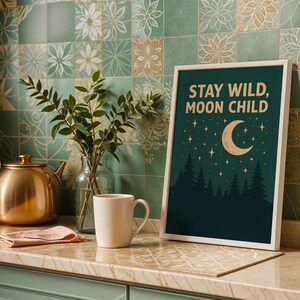 May include: A framed art print with the text "STAY WILD, MOON CHILD" against a dark green background, featuring a crescent moon and stars above a forest silhouette. A white mug and a gold teapot sit on a countertop.