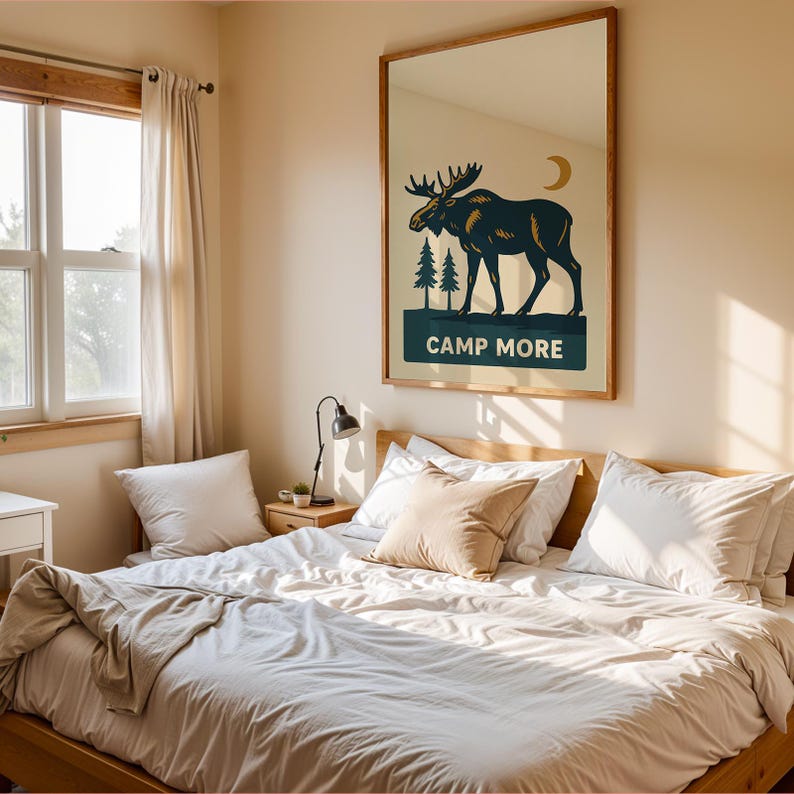 May include: A framed wall art print featuring a moose silhouette with pine trees and a crescent moon. The text "CAMP MORE" is displayed below the moose. The artwork hangs above a bed with white bedding.