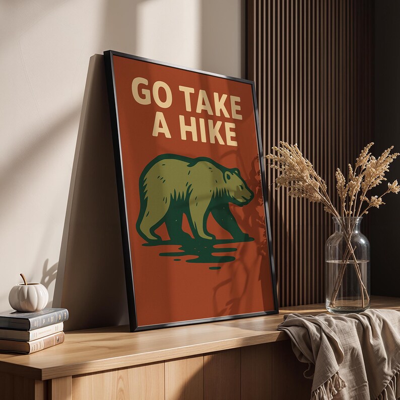 May include: Framed poster with the text "GO TAKE A HIKE" in gold lettering. The poster features a green bear illustration on a rust-coloured background. The bear is walking on a green surface. The poster is in a black frame.