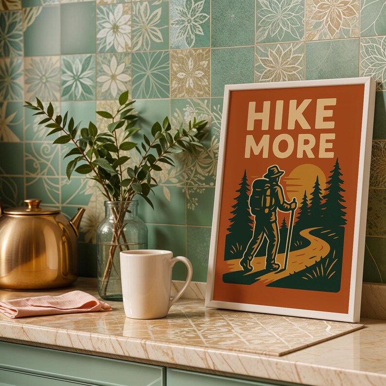 May include: A framed art print with the words "HIKE MORE" in a retro style. The image features a hiker on a trail, with trees and a sunset. A gold teapot, a white mug, and a sprig of greenery are also visible.